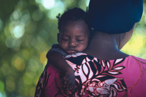 Child resting on caregiver's shoulder in outdoor setting.