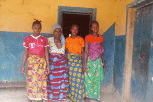 Four women standing outside a yellow and blue house in a rural area.