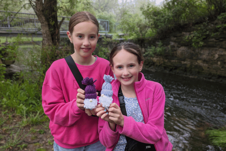 Two girls holding colorful handmade woolen toys outdoors near a stream.