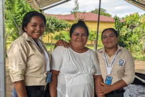Three women smiling outdoors, representing community support and development.
