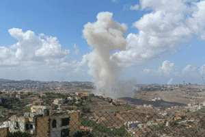 Large explosion with smoke rising over a city with buildings and a fence in the foreground.
