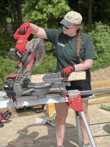 Person operating a mitre saw to cut a wooden board on a construction site outdoors.