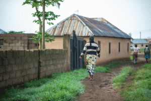 Woman walking towards a rural house with a metal roof and a dirt path.