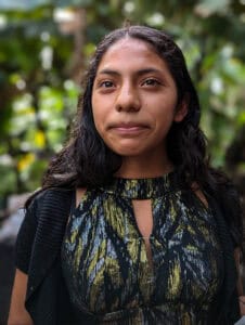 Close-up portrait of a young person standing outdoors with blurred greenery in the background.