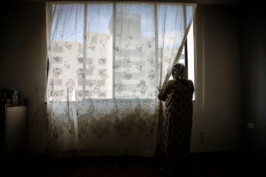 Woman gazing through window with sheer curtains in a dimly lit room.