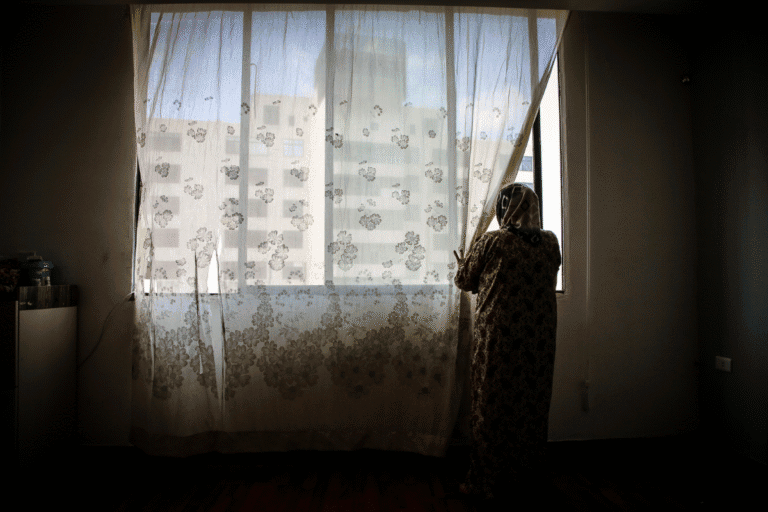 Woman gazing through window with sheer curtains in a dimly lit room.