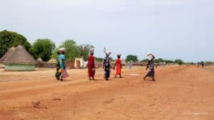 Women walking in a rural village with traditional huts and open landscape.