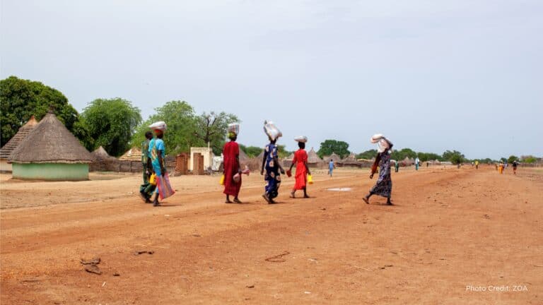 Women walking in a rural village with traditional huts and open landscape.