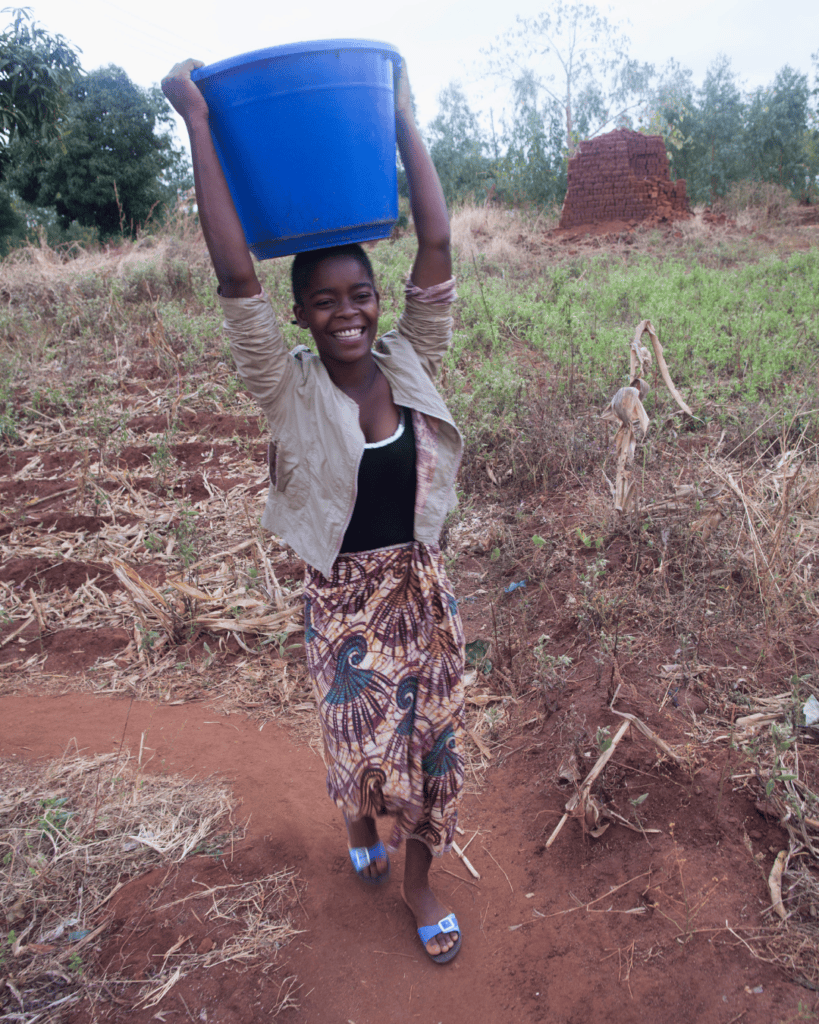 Smiling girl carrying a blue water bucket on her head in a rural area.