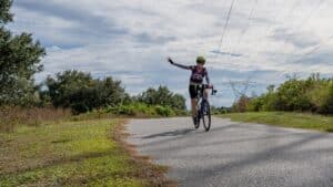 Cyclist riding on a paved trail with one hand raised, surrounded by trees and open sky.