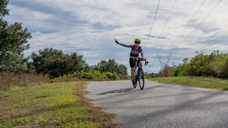 Cyclist riding on a paved trail with one hand raised, surrounded by trees and open sky.