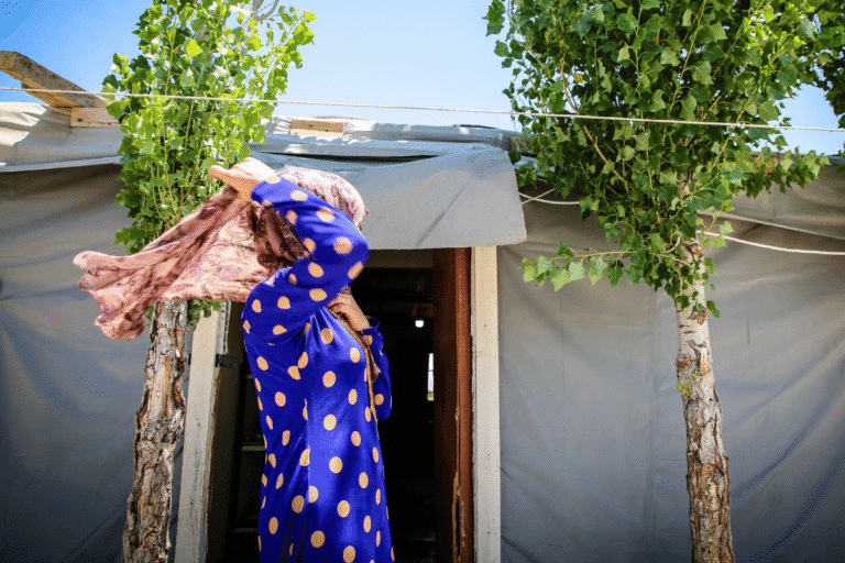 Woman in blue polka dot dress carrying a bag outside a tent in a refugee camp.