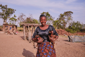 Smiling woman holding chickens in a rural village setting.