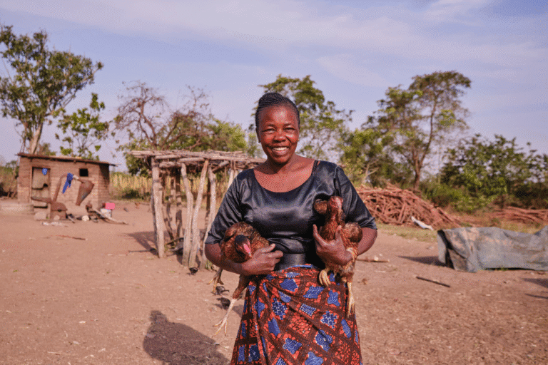 Smiling woman holding chickens in a rural village setting.