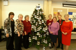 Group of women standing around a decorated Christmas tree with ornaments and a "140" sign.