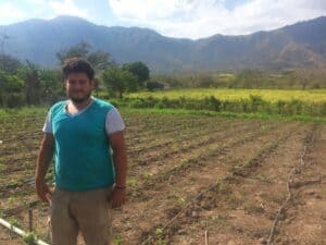 Young man standing in a farm field with mountains in the background.