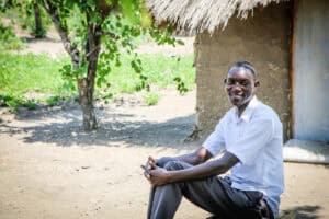 Smiling man sitting outdoors in a rural setting with trees and a thatched hut.