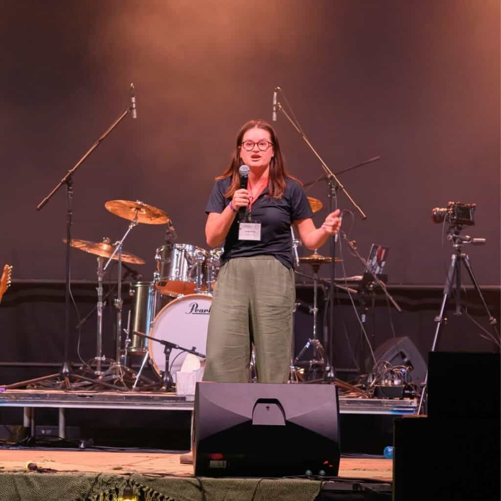 Young woman speaking at AOYC 2026 event on stage with musical instruments.