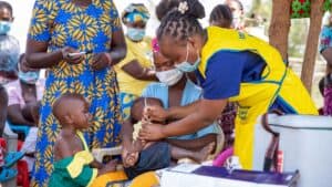 Health worker administering a vaccine to a child seated on a caregiver’s lap in an outdoor clinic setting.