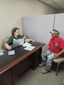 Volunteer with laptop meets a man at a desk to complete paperwork during a disaster assistance visit.