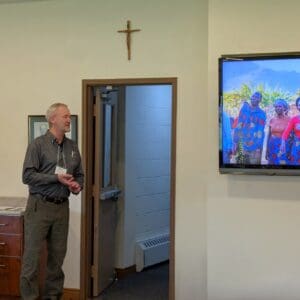 Presenter standing beside a screen displaying a photo while speaking in a meeting room with a cross on the wall.