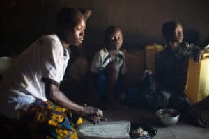 Person grinding grain indoors while two children sit nearby with containers and a bowl in a dimly lit room.