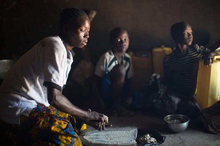 Person grinding grain indoors while two children sit nearby with containers and a bowl in a dimly lit room.