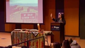 Speaker at a podium presenting to an audience with a screen and table display at Hope College.