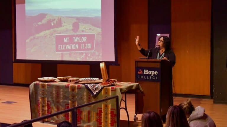 Speaker at a podium presenting to an audience with a screen and table display at Hope College.