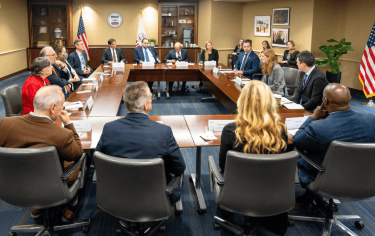 People seated around a conference table during a formal meeting in a government office with flags visible.