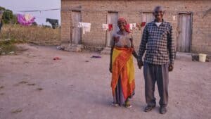 Elderly couple standing outside a mud house in a rural area.