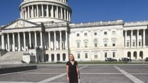 A woman in a black dress stands in the plaza in front of the United States Capitol building under a clear blue sky.