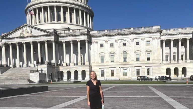 A woman in a black dress stands in the plaza in front of the United States Capitol building under a clear blue sky.