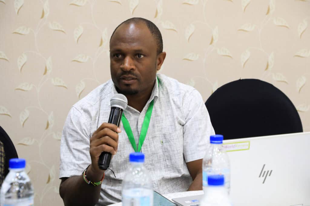 A conference participant speaks into a microphone while seated beside a laptop during a session.