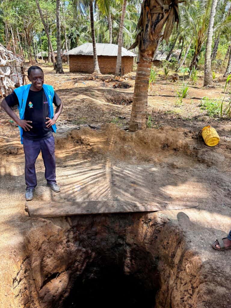 Person stands beside a hand-dug shallow well near homes and palm trees in his community in Kenya.