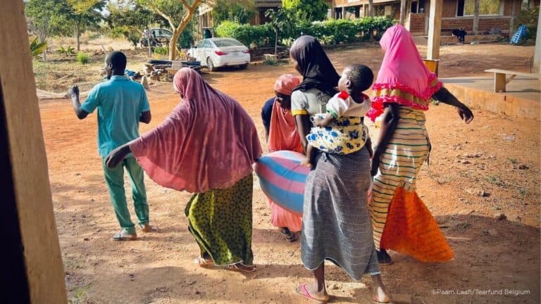 Group of people walking outdoors, some carrying children and belongings near buildings and trees amidst Burkina Faso crisis.