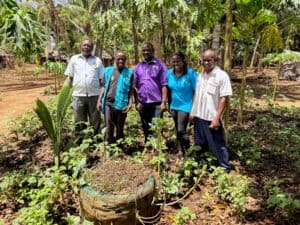 Five people stand in a garden surrounded by vegetable plants and tropical trees in a rural community.