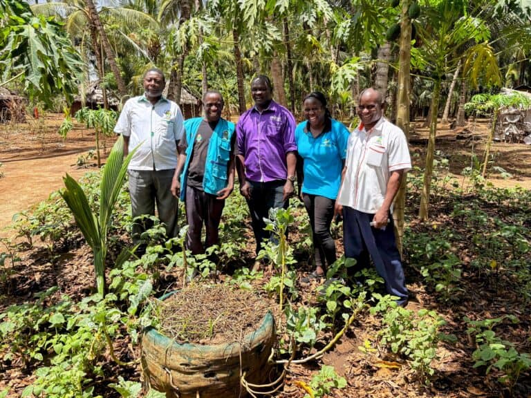 Five people stand in a garden surrounded by vegetable plants and tropical trees in a rural community.