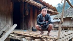 Person crouches on a wooden platform outside home in Nanoy village, Laos. Show more lines