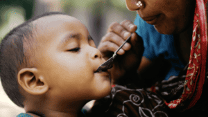 A young child being fed with a spoon while sitting in an adult's arms outdoors.