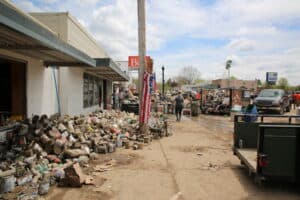 Flood-damaged downtown street with debris piles, cleanup crews, vehicles, and muddy roads after flooding.