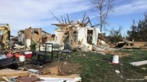 Severely damaged home surrounded by debris after a tornado, with cleanup equipment visible nearby.