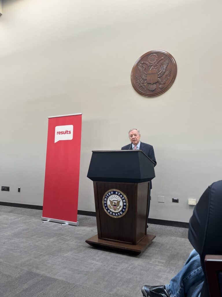 Senator Dick Durbin speaks at a United States Senate podium next to a tall red RESULTS banner.