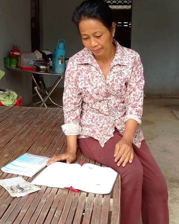 Person seated at a wooden table reading an open notebook inside a home.
