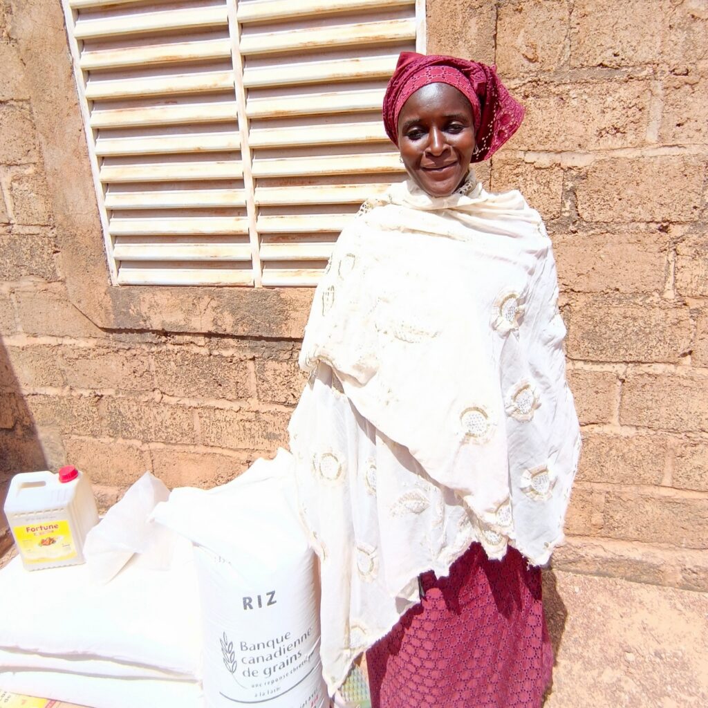 Person standing beside bags of food supplies and a container of cooking oil outside a building.