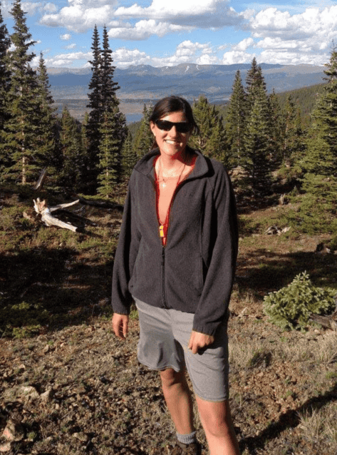 Person standing on a mountain trail with trees and distant peaks in the background.