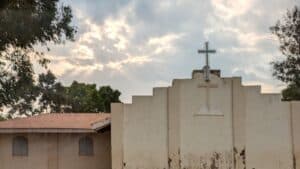 Church building with a cross on the roof beneath a cloudy sky with sunlight breaking through