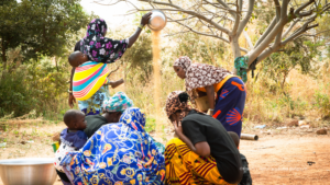 Several adults and children gather outdoors while grain is poured from a metal bowl into a pile.