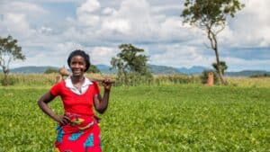 Farmer standing in a field with crops, trees, and distant hills in the background.
