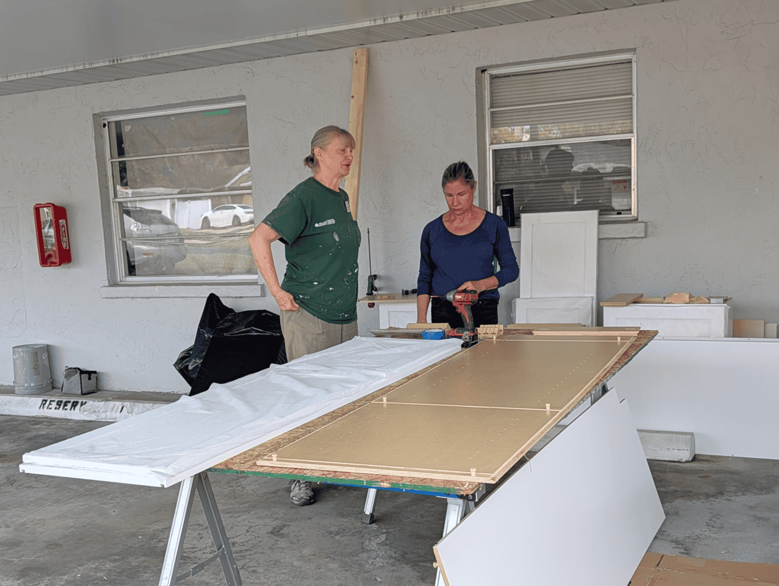 Volunteers prepare cabinet materials and tools for home repairs in a covered outdoor workspace.
